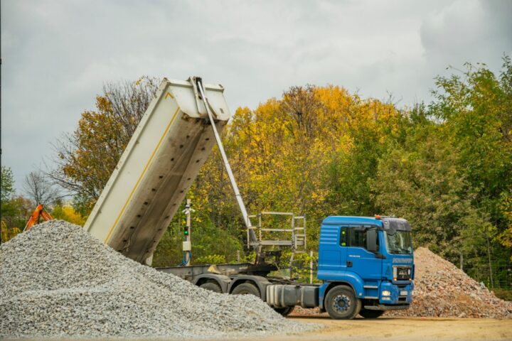 A dump truck tipping a mounded pile of dark screened topsoil onto a gravel driveway beside a new raised bed frame.